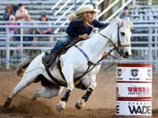 A young rider in a white hat on a grey horse leaning hard into a turn around a red and white barrel, dust kicking up behind