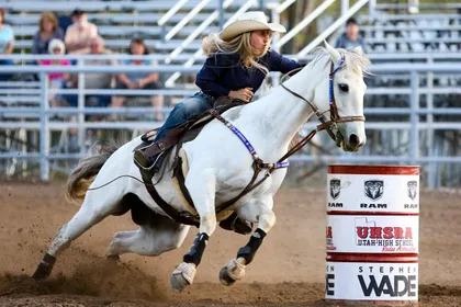 A young rider in a white hat on a grey horse leaning hard into a turn around a red and white barrel, dust kicking up behind
