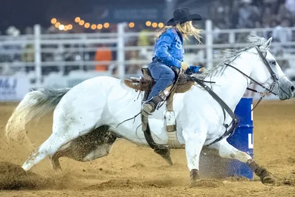 A rider on a grey horse turning tight around a blue barrel under arena floodlights, sand spraying from the horse's feet