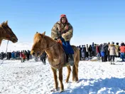 A young rider in a thick fur coat and red hat on a small stocky chestnut horse, standing on trodden snow in front of a crowd of spectators