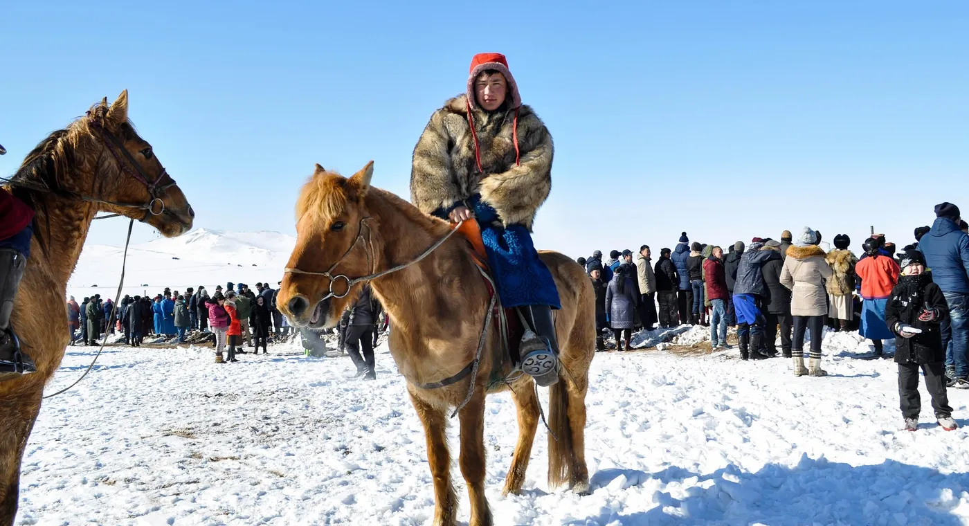 A young rider in a thick fur coat and red hat on a small stocky chestnut horse, standing on trodden snow in front of a crowd of spectators