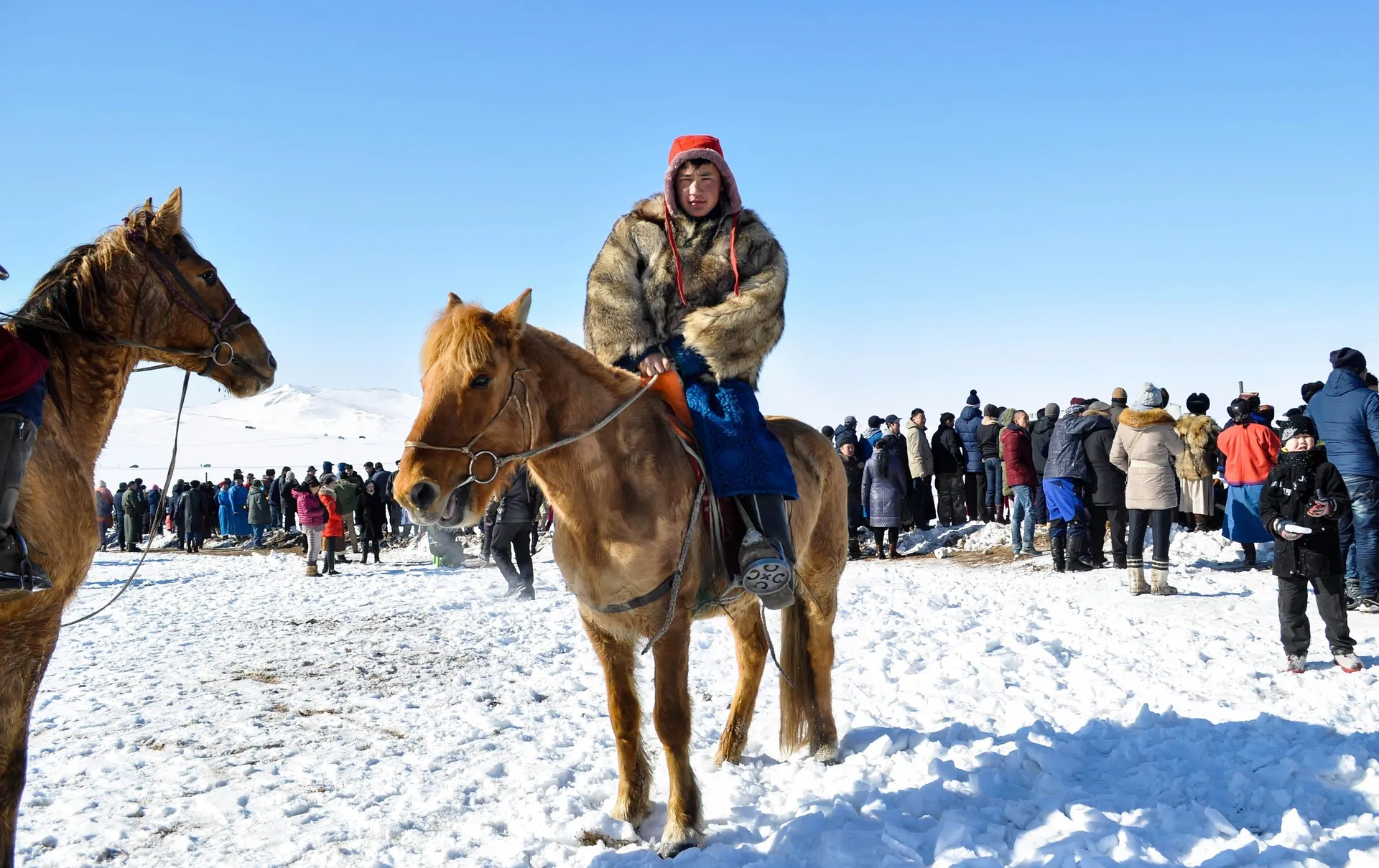 A young Mongolian rider in a thick fur coat and red hat sitting on a small stocky chestnut horse on trodden snow, with a crowd of spectators behind