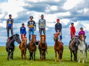 Seven Mongolian boys and young men on small horses on green summer steppe, several of them standing upright on the horses' backs
