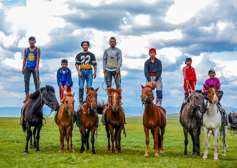 Seven Mongolian boys and young men on small horses on green summer steppe, several of them standing upright on the horses' backs